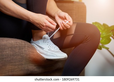 A Close Photo Of The Hands And Legs Of A Sporty Girl In Black Tights Who Is Tying Her Shoelaces On White Sneakers While Sitting On The Couch At Home.