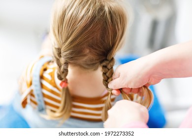 Close up photo of head of toddler girl with blond hair and hands of mum brushing hairs and make plait or pigtail. Mother prepares child for kindergarten or daycare. Family morning routine at bathroom.