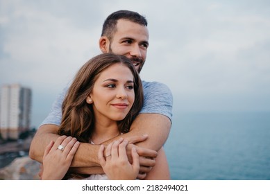 A close photo of a Hispanic man who is hugging from behind his brunette girlfriend in the park in Spain in the evening. A couple of tourists are staring at the sea on a date at the sunset in Valencia. - Shutterstock ID 2182494021
