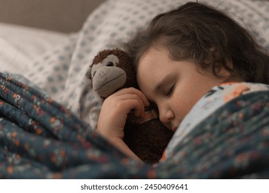 Close up photo of little girl asleep in her bed with a stuffed monkey, peacefully sleeping, very tire, eyes closed with head resting on pillow and snuggle in blankets in her bedroom with soft light - Powered by Shutterstock