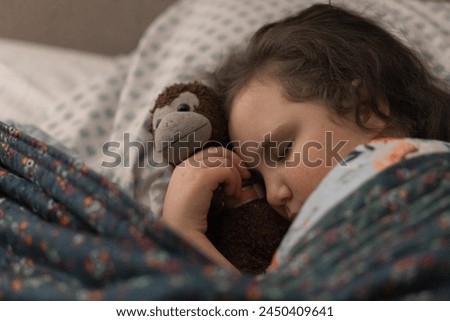 Close up photo of little girl asleep in her bed with a stuffed monkey, peacefully sleeping, very tire, eyes closed with head resting on pillow and snuggle in blankets in her bedroom with soft light