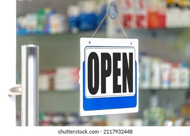 A close up photo of an open sign with white and blue color over blur drug store or Pharmacy background with hard sunlight on a glass door. Backdrop artwork design lifestyle shop text concept ideas.