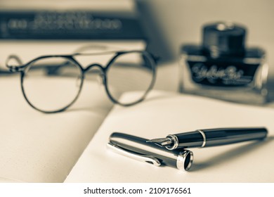 Close up photo of pen,  blurred glasses, book, ink bottle and planner on a gray background. Selective focus. Monochrome photo