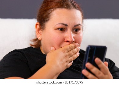 Close up photo of a red hair woman of big size in black t-shirt watching bad news using mobile internet and she looks very awareness, covering her mouth with her hand.