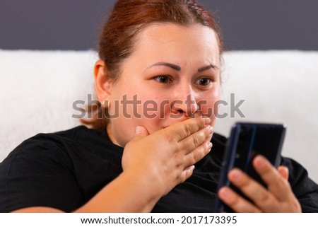 Close up photo of a red hair woman of big size in black t-shirt watching bad news using mobile internet and she looks very awareness, covering her mouth with her hand.