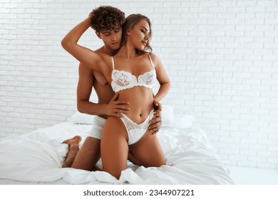 Close up photo of sexy naughty young brunette lady, in seductive nice white underwear, her hot harsh lover is hugging her from the back in bed.