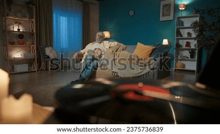 The close up photo shows a vinyl record player with a spinning disc. On the background, an elderly couple is sitting on a sofa. The man looks at the camera and listens to music, and the woman naps