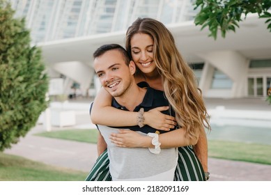 A Close Photo Of A Smiling Couple Of Tourists Who Are Having Fun In The Modern Urban Space On A Date At Sunset In Valencia. A Girl Is Riding Her Boyfriend In The Evening In Spain.