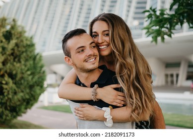 A Close Photo Of A Smiling Couple Of Tourists Who Are Having Fun In The Modern Urban Space On A Date At Sunset In Valencia. A Pretty Girl Is Riding Her Boyfriend In The Evening In Spain.