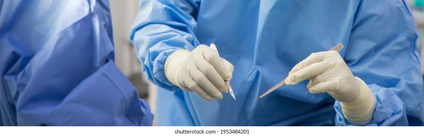 Close Up Photo Of Surgeon 's Hand With Scarpel.Doctor In Blue Surgical Gown Suite Make The Incision With Surgical Knife. Panorama Picture Inside Operating Room.