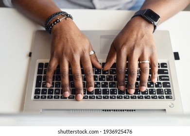 Close up photo of tanned dark male hands with rings, black braided bracelets and a fitness watch, pushing the on the keyboard of the laptop. Top view of the workdesk with modern grey computer