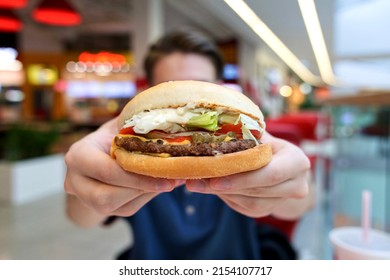 Close up photo of tasty delicious big burger or hamburger, sandwich with beef in man hands at food court in shopping mall. Guy is holding out fast junk food in hands.  - Shutterstock ID 2154107717
