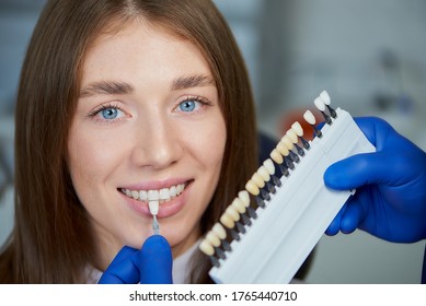 A Close Photo Of A Teeth Shade Sample That Is Matching To A Smiling Female Patient In A Dental Chair. A Doctor Is Checking Teeth Color Samples Before Treatment Procedure In A Dentist's Office.