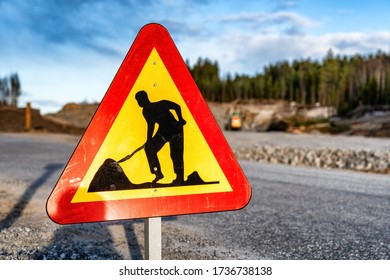 Close Up Photo Of Triangle Formed Red Orange Reflective Warning Traffic Sign - Working Area At Road, Safe, Slow Drive. Blurry Background With Road Construction Area, Forest, Excavator, Evening