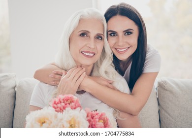 Close up photo of two people white grey hair retired granny hold big bloom blossom peony bunch she get 8-march event daughter hug cuddle sit couch in house indoors