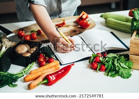Close Up Photo of Woman Hand Writing Recipe in a Empty Notebook at Kitchen Desk with Many Vegetables 