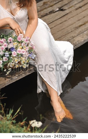 Close up photo of a woman in white summer dress sitting on a wooden bridge with a bouquet of wild flowers in a wicker basket.  Lonely woman on wooden bridge by the lake. Summer season. Space for text