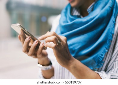 Close up photo of young girl reading messages on a smartphone while standing on urban background. Electronic device in the hands of hipster girl who is posting tweets  by using mobile application.