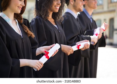 Close Up Photo Of Young Students Dressed In Black Graduation Gown. Campus As A Background. Students Standing In Row, Smiling And Holding Diplomas