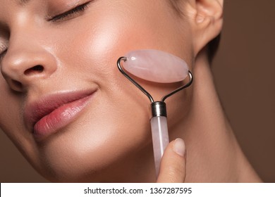 Close up photo of a young woman looking relaxed and smiling while using a natural rose quartz face roller - Shutterstock ID 1367287595