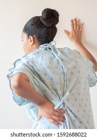 A close up photograph of a middle aged African American mixed race woman with her hand on her lower back or hip hunched over in pain and other hand on the wall wearing a patterned hospital gown.
