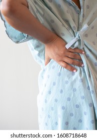 A Close Up Photograph Of A Middle Aged African American Mixed Race Woman With Her Hand On Her Lower Back Or Hip Hunched Over In Pain Wearing A Patterned Hospital Gown.