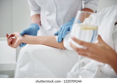 Close Up Of Physician Hand In Sterile Glove Checking Tube And Needle For IV Infusion On Woman Arm. Girl Holding Glass With Lemon Water