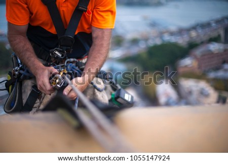 Close up pic of male rope access job industrial worker, using a working safety device descender on static twin ropes abseiling, repairing windows at rise building in  Sydney city CBD, Australia 