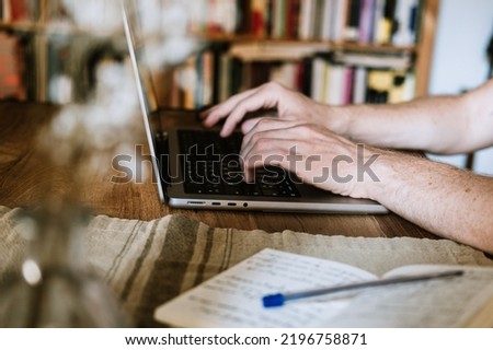 Close up picture of hands typing the laptop at home office. Bookshelf in the background. He has a notepad, pen and a computer. Freelance remote work