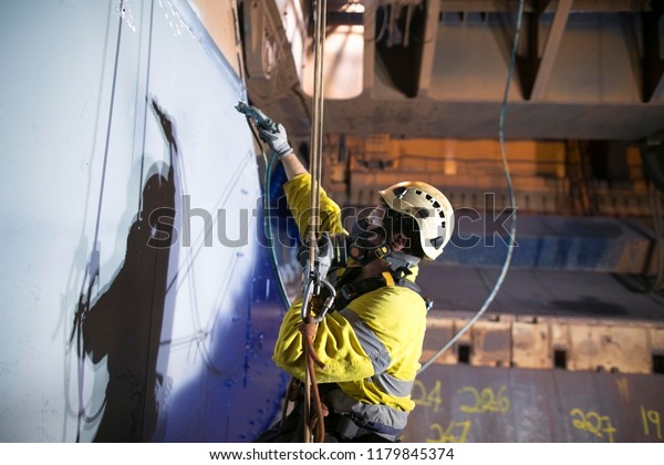 Close up picture of rope access technician\
wearing fully safety harness equipment, respirators for spray\
painting protection descending abseiling commencing spraying blue\
painting job construction