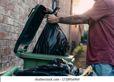 A Close Up Picture Of A Young Male Hand Throwing Rubbish To The Bin At The Street. The Black Rubbish Bin Is For Organic Green Waste Collection. The Bin Lid Is Open. Copy Space. Concept: Ecology