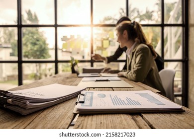 Close Up Piles Of Paper With Blurry Two Asian Business Females Reading Some Documents And Looking At Laptop As Background.