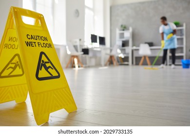 Close Up Plastic Caution Sign With Figure That Slips And Falls Warning Us Of Wet Slippery Office Floor. Black African American Woman Caretaker Housekeeper Cleaning Modern Room Interior In Background