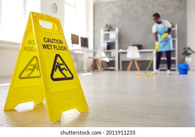 Close Up Of Plastic Sign With Figure That Slips And Falls And Words Caution Wet Floor Black African American Woman Caretaker Housekeeper With Bucket Cleaning Modern Office Room Interior In Background