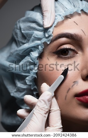 close up of plastic surgeon in latex gloves holding scalpel near woman with marked lines on face isolated on dark grey