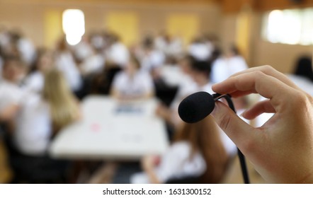 A Close Up Point Of View Image Of A Hand  In Focus Holding  Or Adjusting A Microphone While Public Speaking In Front Of A Deliberately Blurred Audience In A Hall Or Class Room. Announcement Idea