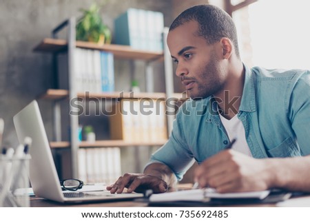 Close up of ponder young african handsome man. He is wearing casual smart, sitting at the workplace, looking in the laptop screen, rewriting the data from it to his report
