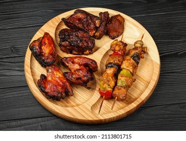 Close Up Portion Of Mixed Grilled Barbecue Combo Plate With Chicken Buffalo Wings, Beef Spare Ribs And Skewer Meat On Wooden Board Over Black Table Planks, High Angle View