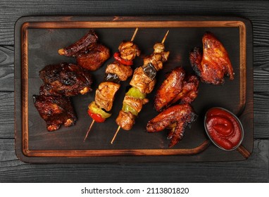 Close Up Portion Of Mixed Grilled Barbecue Combo Plate With Chicken Buffalo Wings, Beef Spare Ribs And Skewer Meat On Wooden Board Over Black Table Planks, Elevated Top View, Directly Above