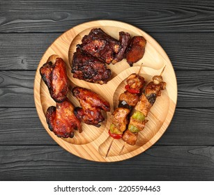 Close Up Portion Of Mixed Grilled Barbecue Combo Plate With Chicken Buffalo Wings, Beef Spare Ribs And Skewer Meat On Wooden Board Over Black Table Planks, Elevated Top View, Directly Above