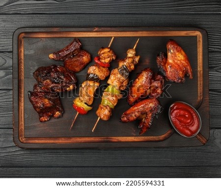Close up portion of mixed grilled barbecue combo plate with chicken buffalo wings, beef spare ribs and skewer meat on wooden board over black table planks, elevated top view, directly above