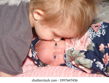 Close Up Portrait Of A 3 Year Old Toddler Kissing His Brand New Baby Sister From Above, Baby Looking At Viewer With Eyes Open, Laying On A Pink Crochet Blanket.