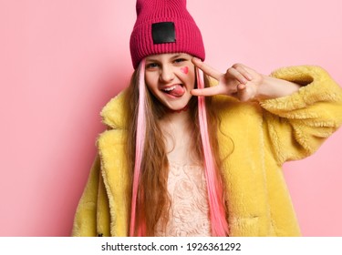 Close up portrait of an active and cheerful teenage girl who is showing V-sign with her fingers and tongue. Little rebel in a pink hat, yellow fur coat posing on a pink background. Adolescence concept - Shutterstock ID 1926361292