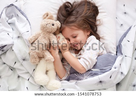 Close up portrait of adorable dark haired little girl calmly sleeping with sweet golden retriever pet in bedroom. Cute elementary age child resting on cozy bedding in nursery hugging her fluffy toys.