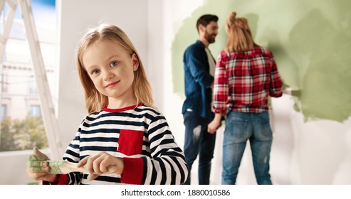 Close Up Portrait Of Adorable Happy Little Cute Girl Smiling To Camera Standing In Room With Paint Brush In Hands. Mom And Dad Painting Wall In Green Colour On Background. Home Repair. Interior Design