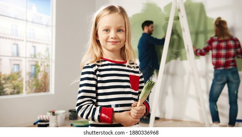 Close Up Portrait Of Adorable Happy Little Cute Girl Smiling To Camera Standing In Room With Paint Brush In Hands. Mom And Dad Painting Wall In Green Colour On Background. Home Repair. Interior Design