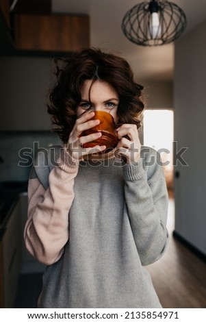 Close up portrait of adorable lovely woman with wavy hairstyle wearing home knitted suit is drinking tea in big cup at home in the kitchen in the morning. High quality photo