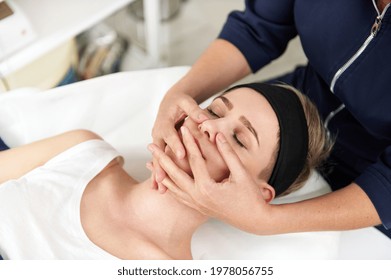 Close Up Portrait Of Aesthetician Hands Doing Professional Rejuvenating Face Massage To Young Blonde Woman Relaxing On Massage Table At Spa Salon. Skin Care Concept