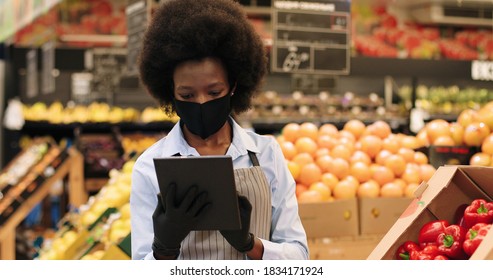 Close Up Portrait Of African Americn Beautiful Female Worker In Black Mask And Gloves Standing In Supermarket And Tapping On Tablet. Woman Manager Browsing On Device In Grocery Shop. Retail Concept