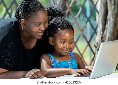 Close Up Portrait Of African Mother And Little Girl With Braided Hairstyle Looking At Laptop. Cute Girl Typing On Keyboard At Table In Garden.